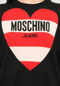 Pull noir avec un grand cœur rayé rouge et blanc affichant le texte "MOSCHINO JEANS" en lettres noires en gras sur la poitrine.