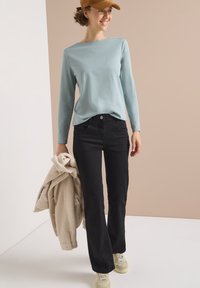 Light blue long-sleeve shirt, black jeans, beige fleece jacket in hand, and yellow sneakers. The background is a neutral tone.