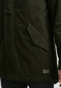 Veste vert foncé avec une finition texturée, présentant une grande poche en biais et une petite étiquette de marque en métal près de l'ourlet.
