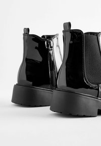 Bottines en cuir verni noir avec des côtés élastiques, un panneau texturé et une semelle en caoutchouc épaisse et basse. Fermeture zippée à l'arrière.