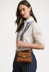 Brown textured handbag with a gold logo, showcasing a structured design and short handle, carried alongside a white shirt and pinstriped pants.