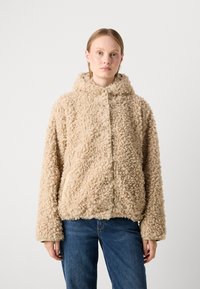 Kvinne iført en beige krøllete fleecjakke med hette og blå jeans, stående mot en helt lys bakgrunn.