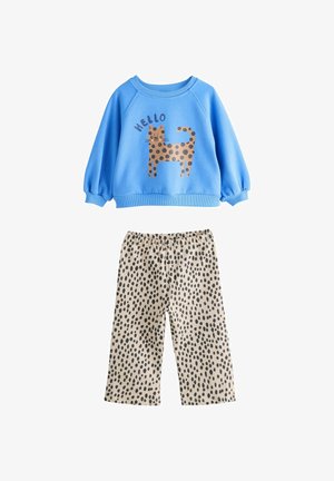 Sweat-shirt bleu avec un graphique d'un chat tacheté marron et du texte disant "BONJOUR," associé à un pantalon beige avec des motifs de points noirs.