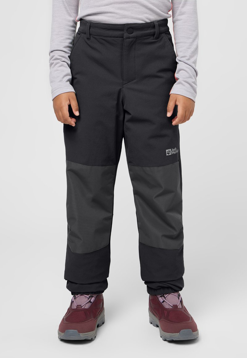 Bambino che indossa pantaloni da esterno neri e grigio scuro con polsini elastici e scarpe da trekking color bordeaux su sfondo chiaro.