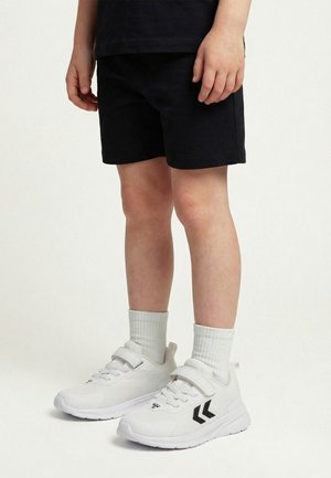 Kind draagt zwarte korte broek, witte hoge sokken en witte sneakers met zwarte chevron-logo's, staand op een lichte achtergrond.