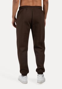 Mann trägt dunkelbraune Jogginghose und weiße Nike Air Sneaker, steht mit entspannten Armen an den Seiten, Rückansicht vor einfarbigem Hintergrund.