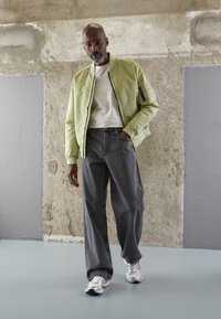 Light green bomber jacket over a white shirt, paired with loose gray jeans and shiny silver sneakers. Concrete backdrop with a smooth floor.
