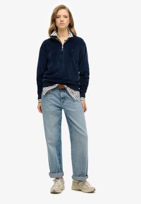 Sudadera de forro polar azul marino con cuello con cremallera, llevada sobre una camisa a rayas. Unos vaqueros azul claro y zapatillas beige completan el conjunto.