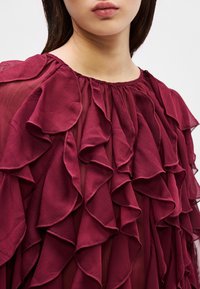 Ruffled burgundy blouse with a lightweight, sheer fabric. Features multiple layered ruffles along the neckline and sleeves with a textured finish.