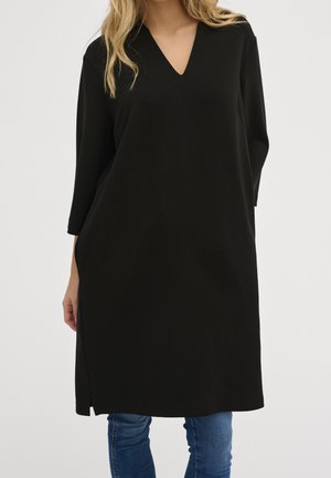 Femme portant une robe-tunique noire à manches longues avec un col en V, associée à un jean bleu, debout devant un fond clair uni.
