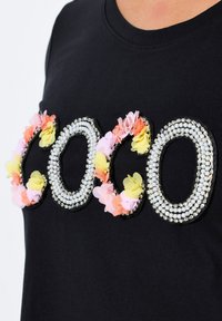 Camiseta negra con la aplicación "COCO" hecha de perlas y flores de tela coloridas en rosa, amarillo y naranja. Manga corta, cuello redondo.