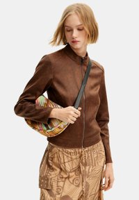 Braune Kunstlederjacke mit Reißverschluss vorne, hohem Kragen und langen Ärmeln, kombiniert mit einer mehrfarbigen geflochtenen Tasche und gemusterten beigen Hosen.