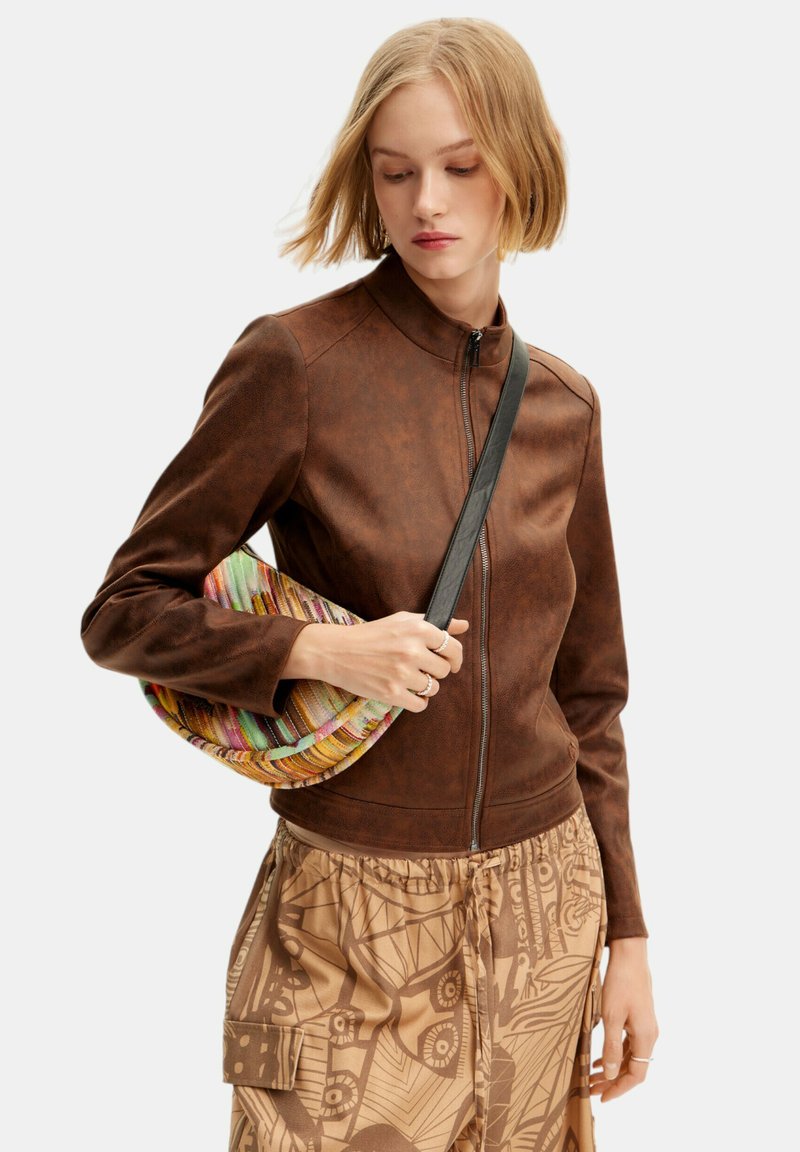 Braune Kunstlederjacke mit Reißverschluss vorne, hohem Kragen und langen Ärmeln, kombiniert mit einer mehrfarbigen geflochtenen Tasche und gemusterten beigen Hosen.
