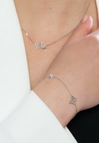 Srebrny naszyjnik i bransoletka w kształcie motyla, cechujące się minimalistycznym wzornictwem z otwartymi i solidnymi wzorami, noszone na skórze na neutralnym tle.