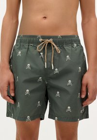 Shorts de baie verzi cu un model repetat de craniu și oase încrucișate, talie elastică cu șnur bej și buzunare laterale.