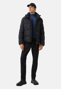 Mann in schwarzer Mütze, schwarzer Steppjacke, blauer Zip-Up-Jacke, schwarzen Jeans und schwarzen Schuhen, der vor einem weißen Hintergrund steht.