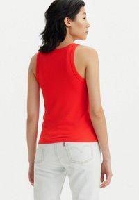 Top de tirantes rojo con un diseño ajustado. La modelo lo lleva con unos jeans de tiro alto de color azul claro que cuentan con bolsillos traseros y detalles de costura.