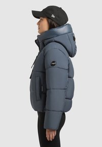 Marineblauwe pufferjack met kap, gequilt ontwerp, rits aan de voorkant, logo patch op de mouw en een elastische zoom. Gecombineerd met een zwarte pet.