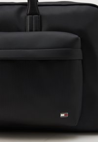 Zwarte leren tas met een glad bovenste gedeelte, een geribbelde voorkant en een klein logo accent in de rechteronderhoek.