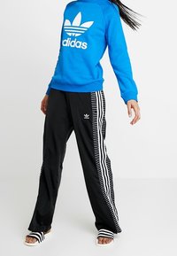 Modrá mikina s bílým logem Adidas, černé sportovní kalhoty s bílými pruhy a vzorem a černobílé pruhované sandály.