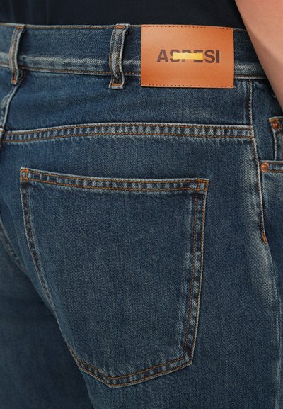 Bakifrån av en person som bär blå denimjeans med synlig bakficka och en brun läderlogotyp på midjebandet.