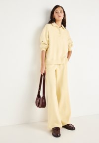 Κίτρινο φλις πόλο με γιακά και τρία κουμπιά, συνδυασμένο με παντελόνι με φαρδύ πόδι. Τα αξεσουάρ περιλαμβάνουν μια μικρή μπορντό τσάντα και λόφερ.