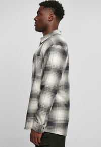 Flannel skjorte i sort og hvid plaid, med knap-ned krave, lange ærmer og en enkelt brystlomme. Blød stofstruktur er synlig.