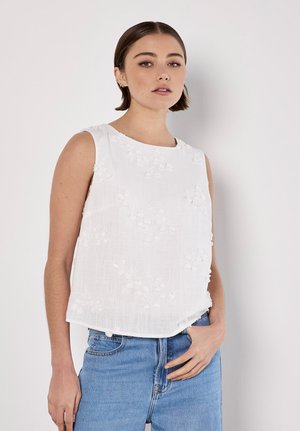 Junge Frau mit kurzen braunen Haaren trägt ein ärmelloses weißes, blumengemustertes Oberteil und blaue Jeans, steht vor weißem Hintergrund.