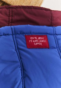 Kołnierz niebiesko-bordowej kurtki puchowej z czerwoną metką z napisem „Zaraz sprawisz, że magia się wydarzy” białym tekstem.