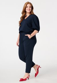 Haut en V texturé bleu marine foncé avec des manches trois quarts associé à un pantalon ajusté assorti. Escarpins à talons ouverts en vernis rouge avec détail en nœud.