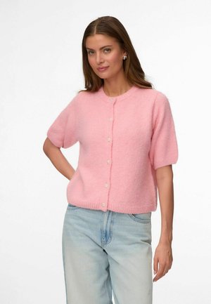 Vrouw met lang bruin haar, die een roze kort mouw gebreid vestje met knopen draagt en lichtblauwe spijkerbroek, staand tegen een effen witte achtergrond.