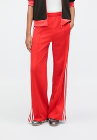 Czerwone szerokie spodnie dresowe z białymi paskami po bokach, elastycznym paskiem w talii i lekkim materiałem, stylizowane z czarną kurtką i szarymi butami.