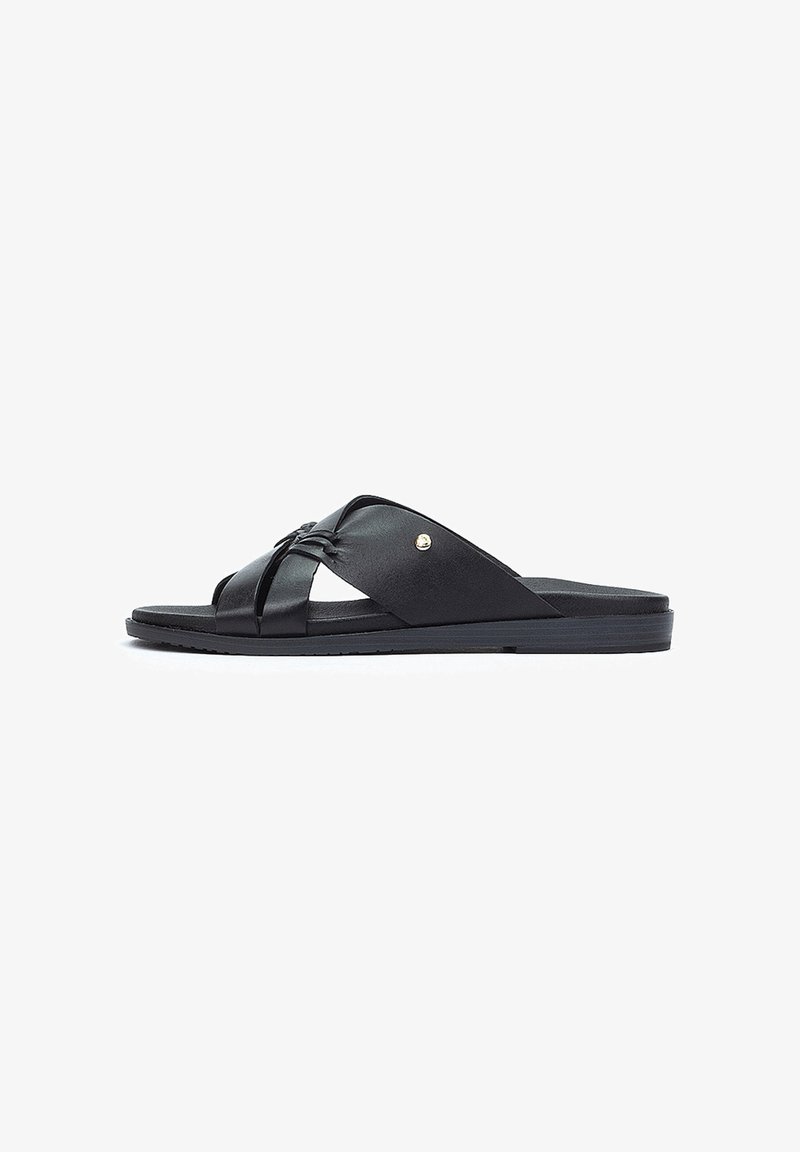 Des sandales à glisser en cuir noir avec des brides croisées et un petit accent métallique. Semelle plate avec un fond texturé pour une bonne adhérence.