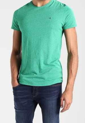 Mann in einem taillierten grünen Kurzarm-T-Shirt und dunkelblauen Jeans, der mit einer Hand in der Tasche vor einem schlichten Hintergrund steht.