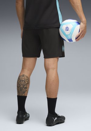 Parte inferior del cuerpo de un jugador de fútbol con pantalones cortos negros y botas de fútbol, sosteniendo un balón blanco y azul, con un tatuaje detallado en la pantorrilla izquierda.