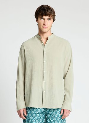 Jeune homme portant une chemise beige clair boutonnée avec un col mandarin et un short bleu à motifs avec des dessins de tortues blanches.