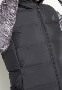 Chaleco acolchado negro de Adidas llevado sobre una camiseta de manga larga gris con patrón, cremallera cerrada, torso y parte del rostro visibles.