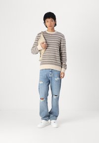 Suéter de punto en color marrón claro con patrones geométricos oscuros, combinado con unos vaqueros desgastados de color azul claro y zapatillas blancas. Bandolera beige colgada del hombro.