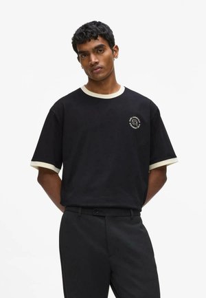 Jeune homme aux cheveux courts et bouclés porte un T-shirt noir avec bordure blanche et un pantalon noir, debout, les mains derrière le dos.