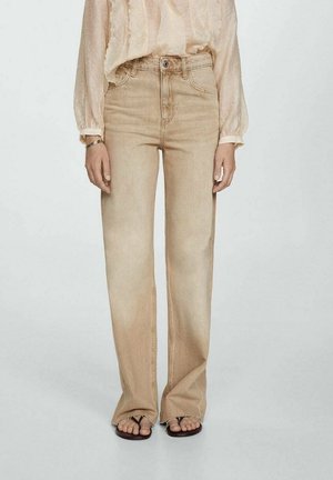 Kvinde iført beige højlivsvidde jeans, let gennemsigtig langærmet bluse og sorte sandaler, stående mod en ensfarvet baggrund.