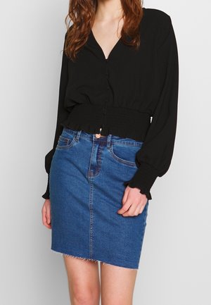 Mujer que lleva una blusa negra de manga larga con botones, cintura y puños fruncidos, combinada con una minifalda vaquera azul.