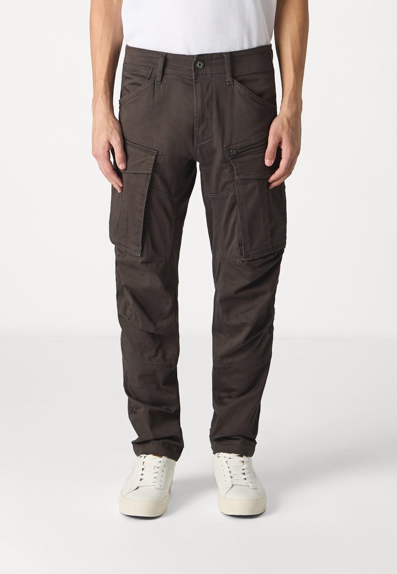 Homme portant un pantalon cargo marron foncé avec de grandes poches latérales et des baskets blanches, debout devant un fond blanc uni.