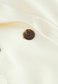 Cremefarbener Stoff mit glatter Textur, ausgestattet mit einem dunkelbraunen, runden Knopf mit vier Löchern und leicht erhabenen Stickdetails.