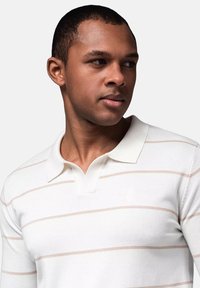 Witte polo met lange mouwen en lichtbeige horizontale strepen, zachte textuur en een klassieke kraag. Bevat een subtiele logo op de borst.