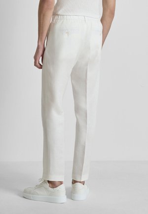 Persoon draagt witte op maat gemaakte broek met achterzakken en witte sneakers, staand tegen een effen lichtgrijze achtergrond.