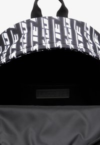 Sac à dos noir avec un motif de logo blanc en répétition, texture lisse, et un compartiment principal zippé avec une étiquette de marque à l'intérieur.