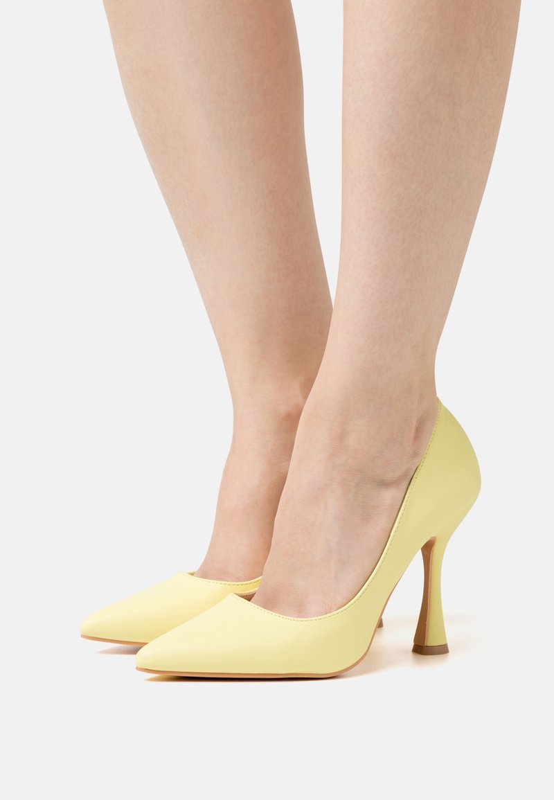 Jambes portant des escarpins jaune pâle à bout pointu avec des talons aiguilles fins et évasés sur fond blanc.
