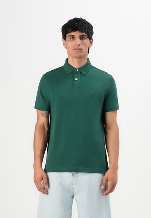 Homme portant un polo vert foncé avec un petit logo sur la poitrine et un jean bleu clair, se tenant devant un fond blanc.