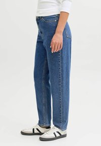 Augsta vidukļa zilas džinsa bikses ar taisnu kāju dizainu un klasisku piecu kabatu stilu. Valkātas kopā ar baltām un melnām sporta kurpēm.