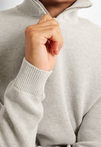 Hellgrauer gestrickter Fleecepullover mit Reißverschluss, hohem Kragen und gerippten Bündchen. Der Stoff wirkt weich mit einem strukturierten Muster und dezenten Details.